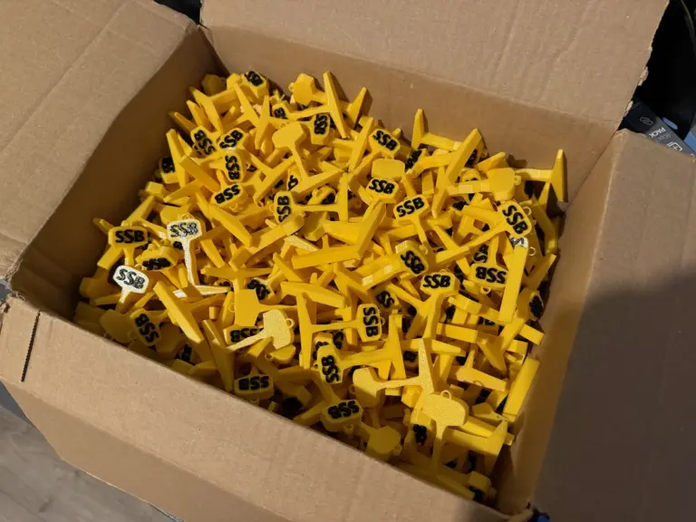Karton voller gelber 3D-gedruckter Schlüsselanhänger in Form eines Schienenquerschnitts mit schwarzem Logo